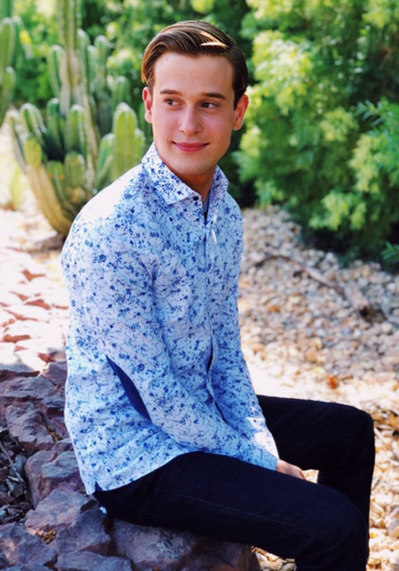 Tyler Henry in light blue pattern top and black jeans sittin go on a rock outside